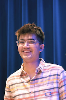 A young man in glasses smiling confidently as he stands in front of a microphone.