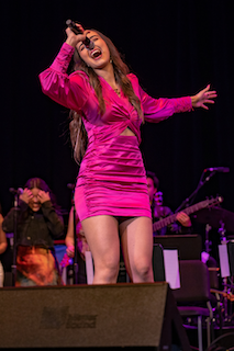 A woman in a pink dress captivatingly sings on stage, captivating the audience with her performance.