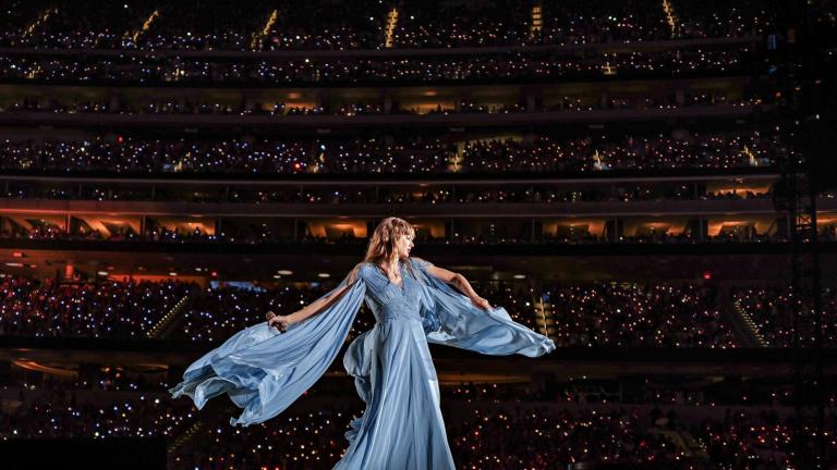 Taylor Swift in a flowy dress on stage with a backdrop of a packed stadium of fans