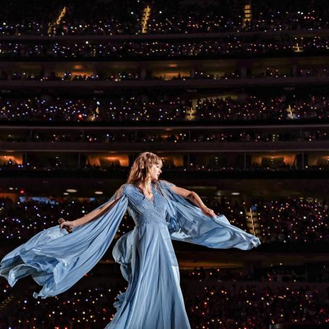 Taylor Swift in a flowy dress on stage with a backdrop of a packed stadium of fans