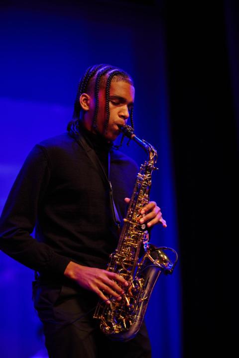 A young man wearing a black shirt is playing a saxophone on a dimly lit stage